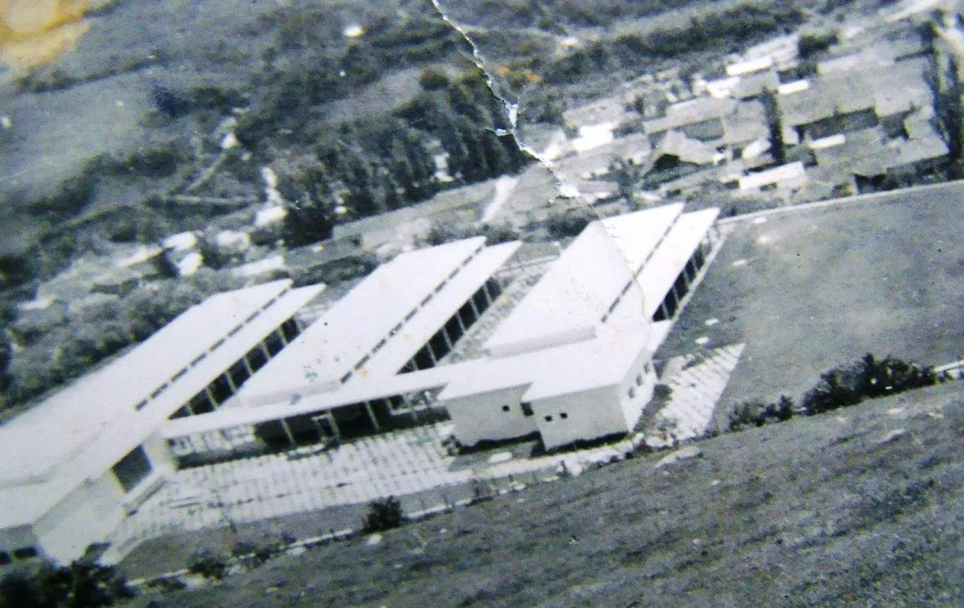 Fotografía antigua desde lo alto: los pabellones blancos de la sede inaugurada en 1954, con el pueblo detrás.
