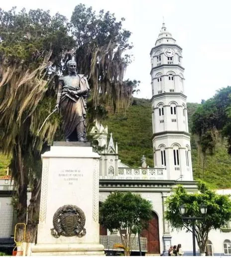 La estatua pedestre del Libertador sobre su pedestal en la Plaza Bolívar, con la torre de la basílica al fondo.