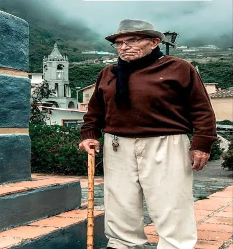 Un hombre mayor, de sombrero, chaqueta y bastón, de pie junto al monumento de la plaza; detrás, la torre de la iglesia y el páramo entre nubes.