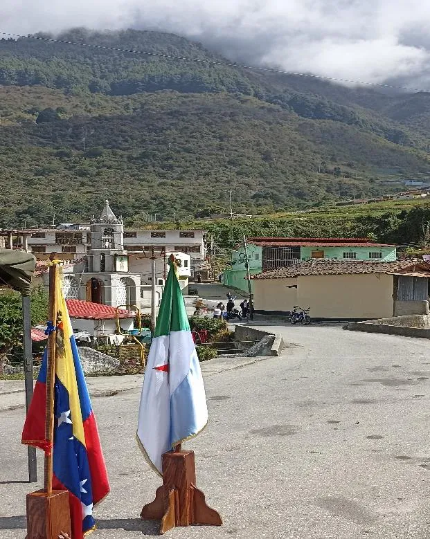 Dos banderas en el atrio: la de Venezuela y la parroquial, con la iglesia y las casas de La Venta al fondo.