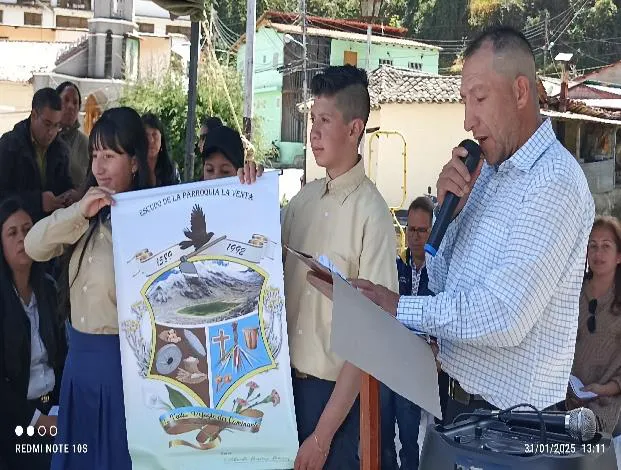Presentación del escudo parroquial ante el público, sostenido por dos estudiantes mientras un hombre lee ante el micrófono.