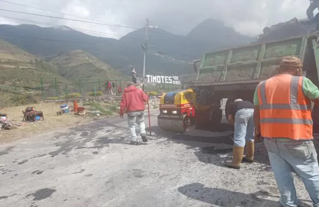 Cuadrilla de obreros asfaltando la vía a la entrada de Timotes, con el camión volteo descargando y el cartel del pueblo al fondo.