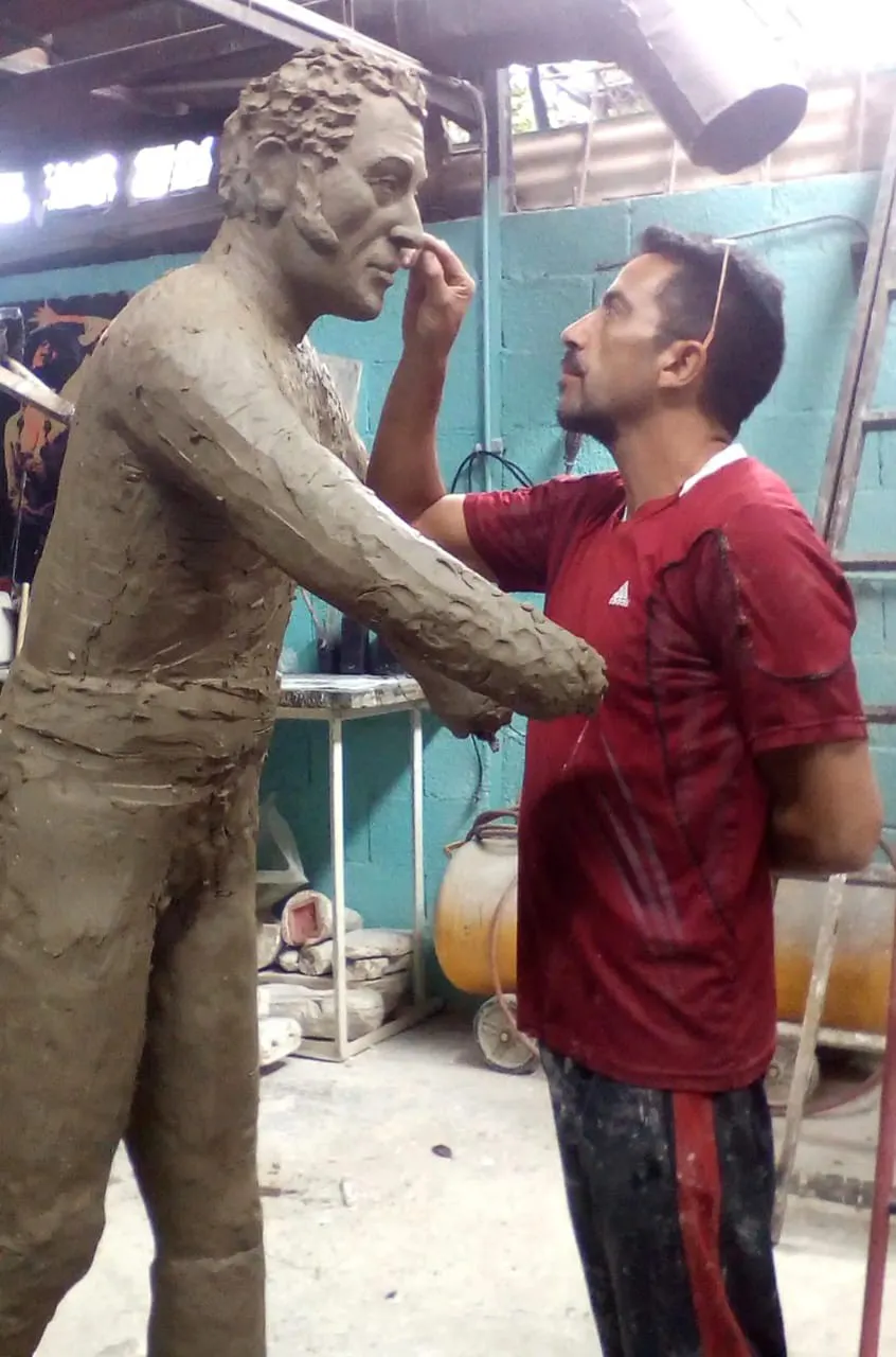 Un escultor en su taller, de franela roja, retoca con los dedos el rostro de una figura de barro de tamaño natural.