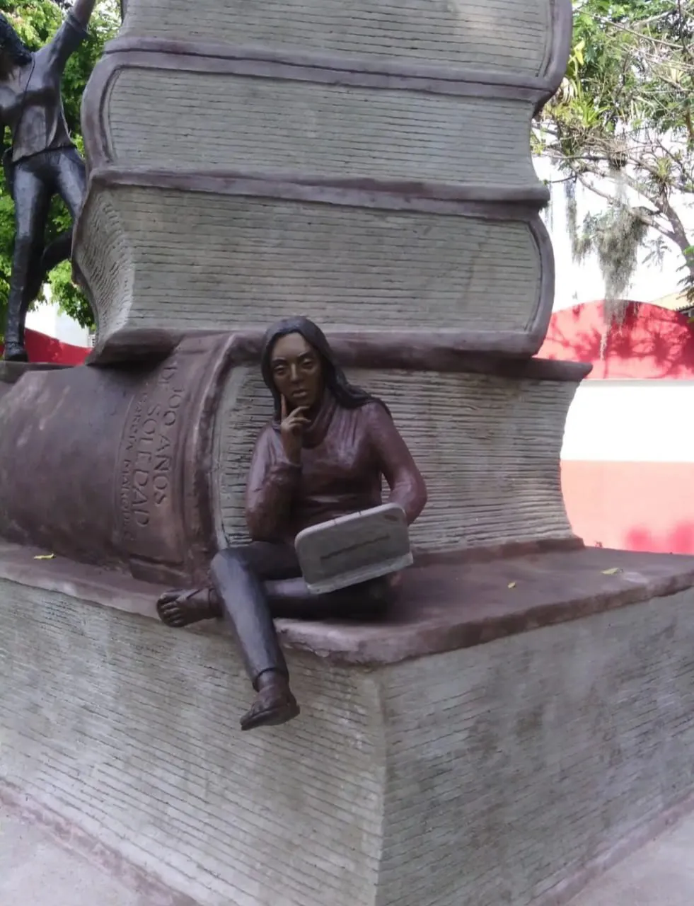 Conjunto escultórico de libros gigantes apilados, con una estudiante sentada en la base leyendo con la mano en el mentón.