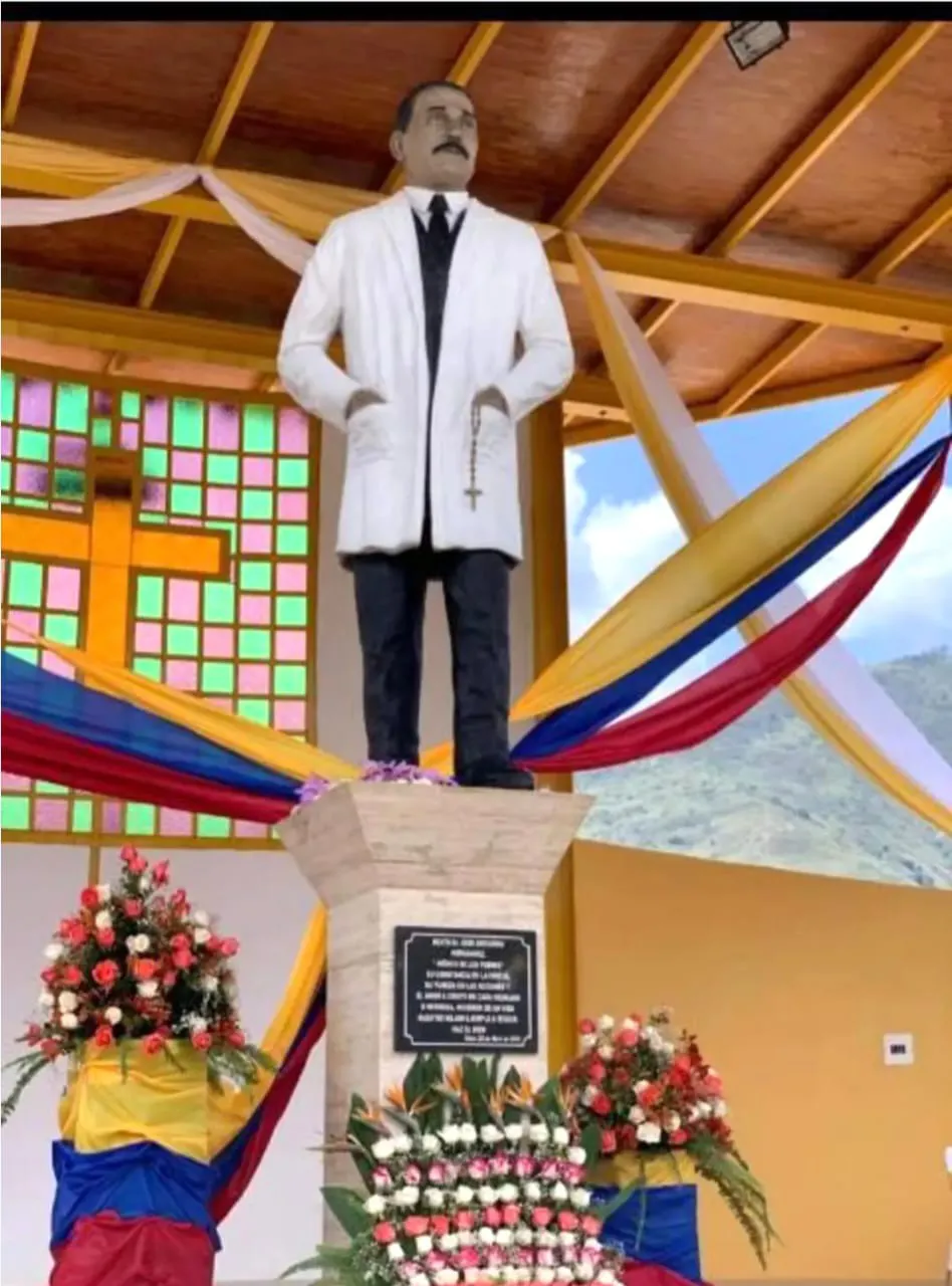 Estatua de José Gregorio Hernández con su bata blanca, sobre un pedestal con placa y rodeada de flores y cintas tricolores.
