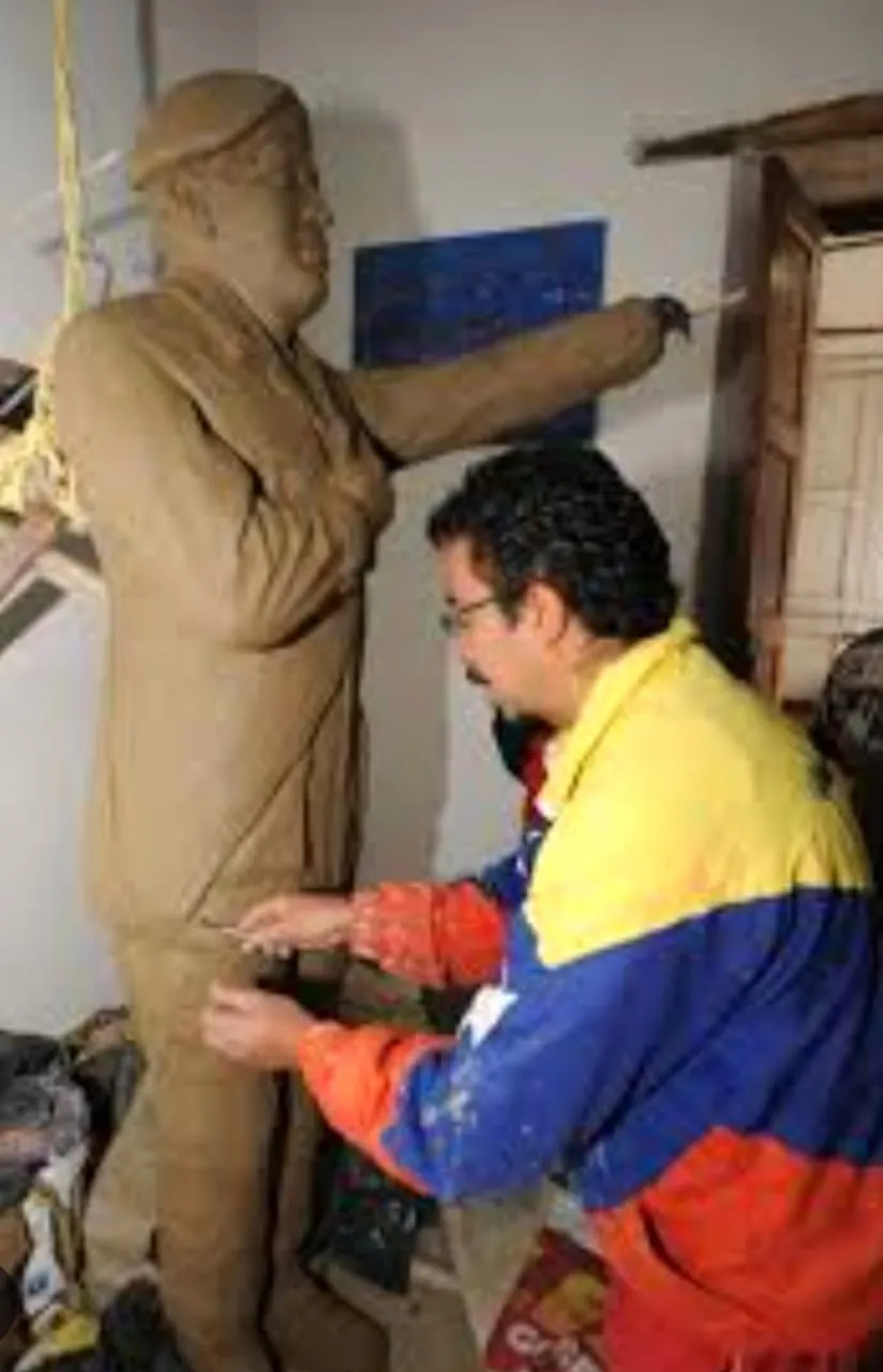 El escultor, con chaqueta tricolor, modela en barro una figura de hombre con gorra.