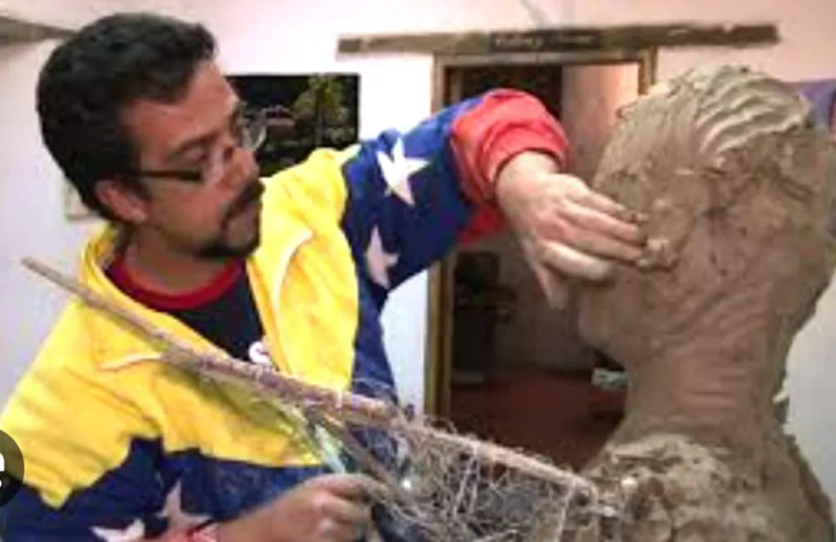 El escultor, con chaqueta tricolor, modela la cabeza de un busto de barro.