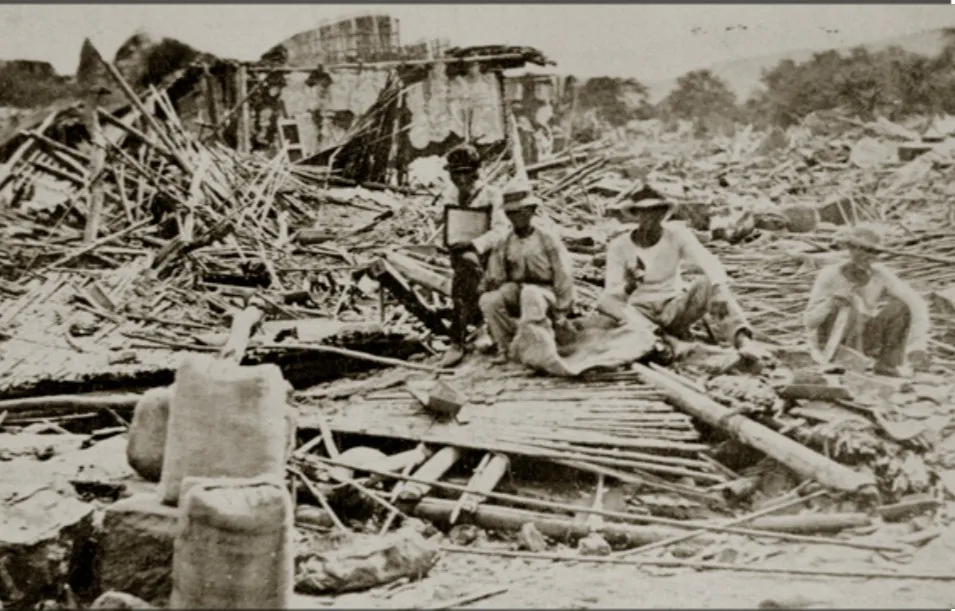 Fotografía antigua: unos hombres sentados entre escombros de maderas y tejas derrumbadas.