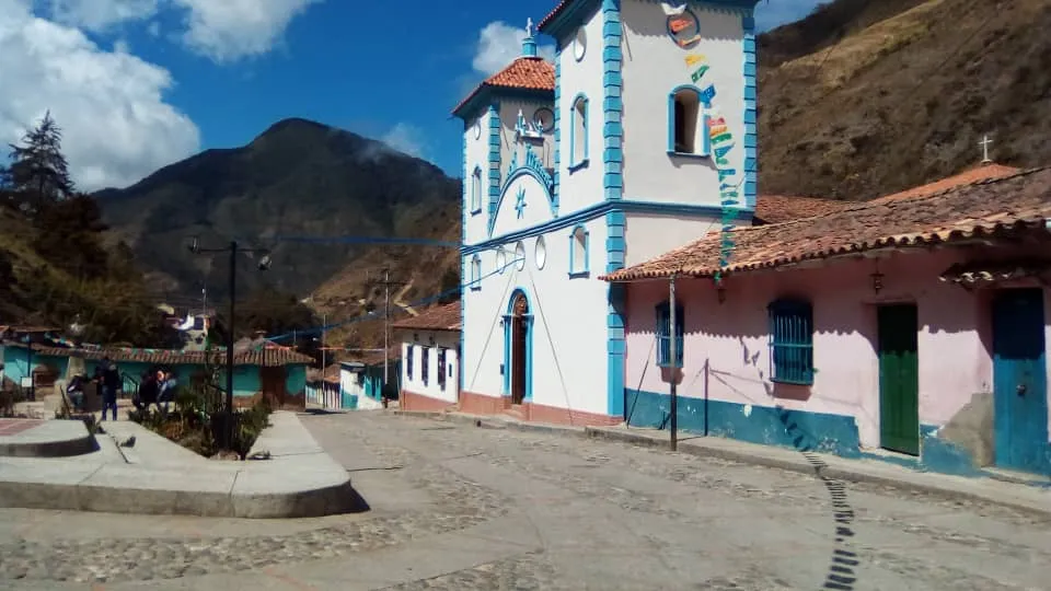 Una iglesia blanca de torre única con ribetes azules, frente a la calle y entre montañas.