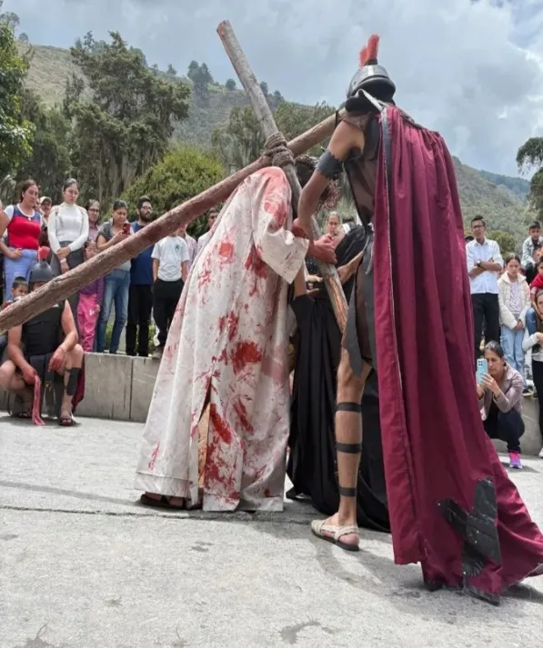 Viacrucis viviente: el Nazareno, con la túnica manchada de sangre, carga la cruz junto a un soldado romano de capa roja, ante el público.