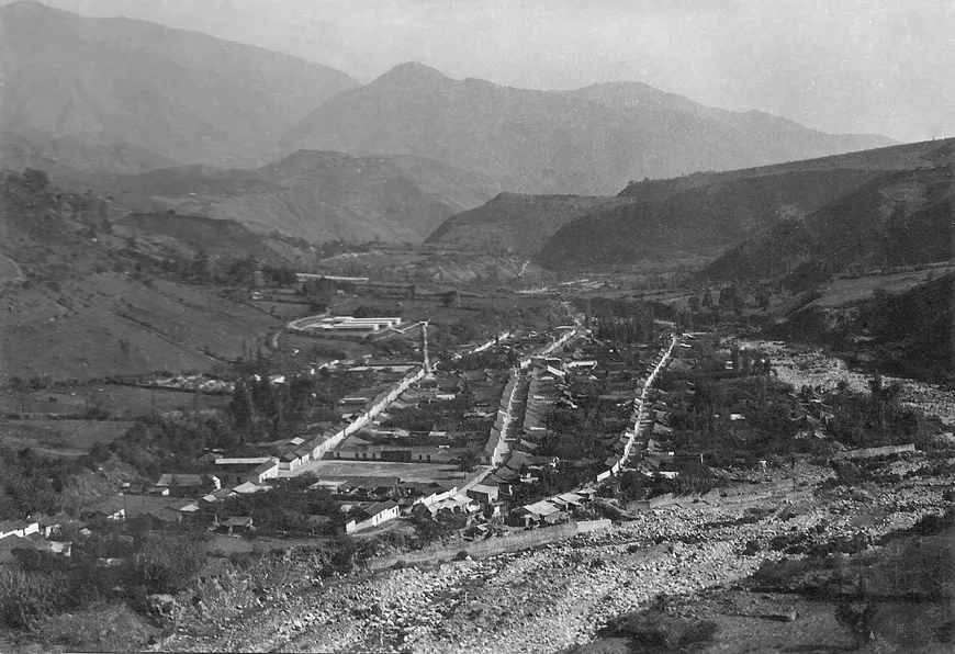 Vista panorámica en blanco y negro del pueblo en el valle, rodeado de montañas.