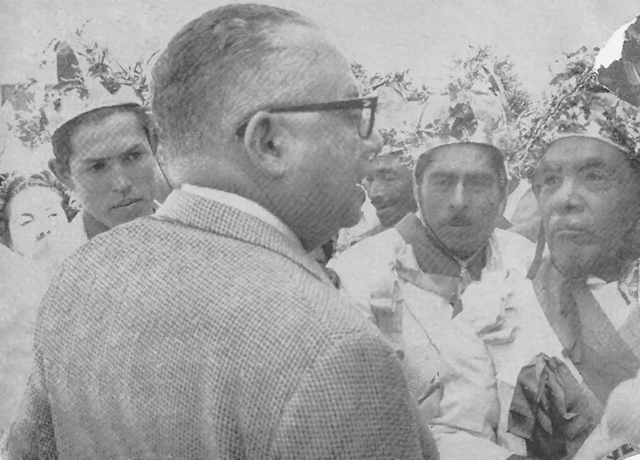 Fotografía antigua de un hombre de lentes rodeado por danzantes con tocados de flores.