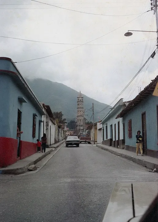 Fotografía en color de una calle recta con casas de fachadas blancas y la torre de la iglesia al fondo.