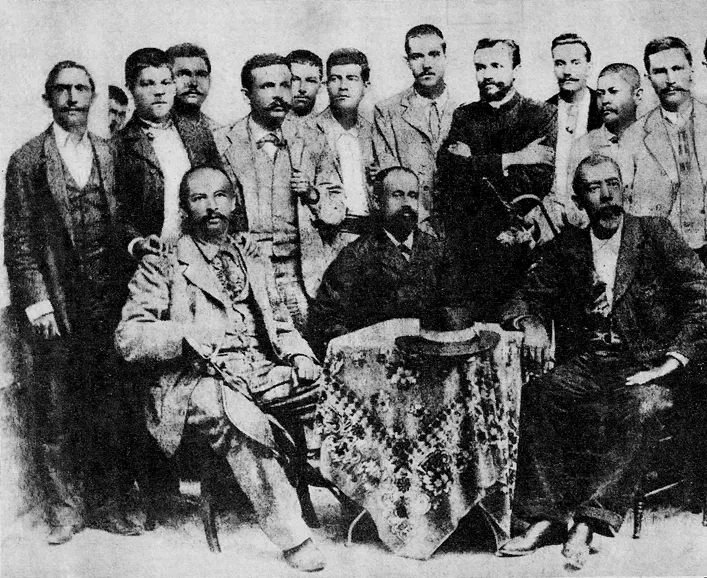 Fotografía antigua de un grupo numeroso de hombres, de pie y sentados alrededor de una mesa con mantel.