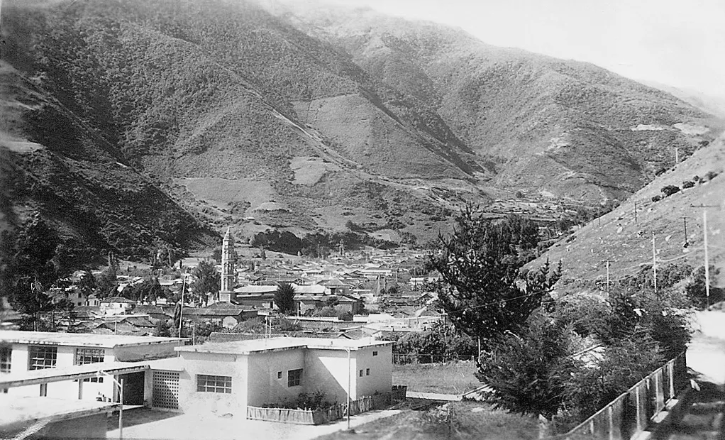 Vista en blanco y negro del valle, con casas en primer plano y las montañas al fondo.
