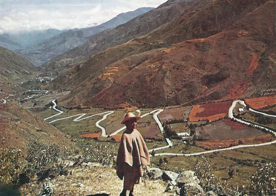 Fotografía en color de un camino que baja en zigzag por una cañada entre montañas; en primer plano, una persona con ruana.