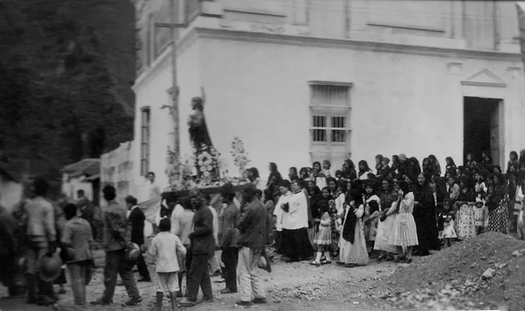 Fotografía antigua de una procesión: una multitud frente a una casa blanca de dos pisos.