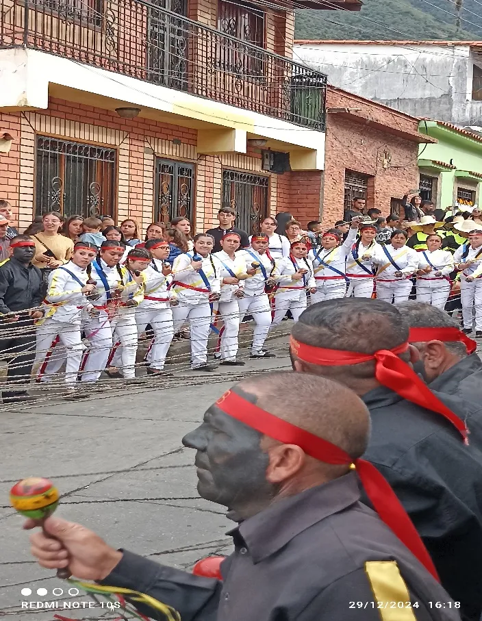 Los Negros de San Benito, con el rostro tiznado y la vincha roja, en primer plano con sus maracas, mientras la fila de danzantes de blanco tensa los cordeles calle abajo.