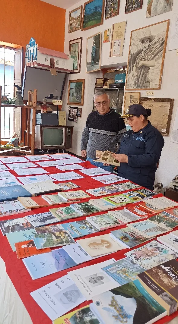 Mesa con la exposición de las publicaciones timotenses, y al fondo la pared de cuadros y retratos; el cronista acompaña a una visitante que hojea uno de los libros.