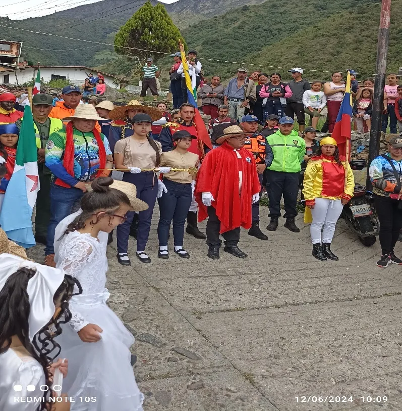 Acto cívico en la calle del páramo: portaestandartes, un caracterizado de Bolívar con capa roja, niñas de traje blanco y los vecinos alrededor.