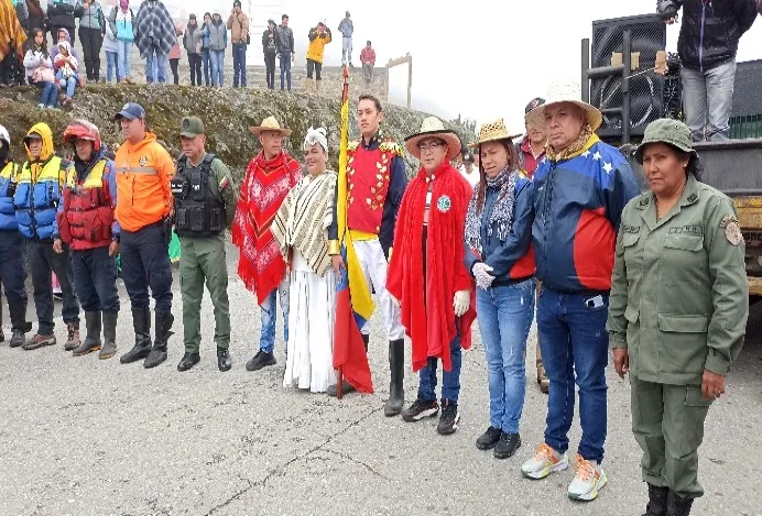 Formación de autoridades, protección civil y vecinos en un acto cívico, con un caracterizado de Bolívar sosteniendo la bandera y dos personas de traje regional.