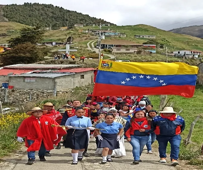 Escolares de uniforme y ruana roja suben por el camino del páramo detrás de una gran bandera nacional, en la conmemoración del paso del Libertador.