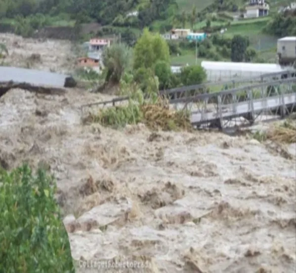 La crecida arrastra troncos y ramas contra el puente metálico, con las casas y los invernaderos en la orilla.