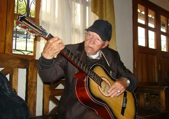 Un anciano de gorra afina y toca su guitarra, sentado en el corredor de una casa.