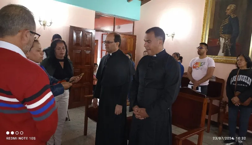 Dos sacerdotes de sotana, de pie ante los vecinos en el salón del Concejo, bajo el retrato del Libertador.