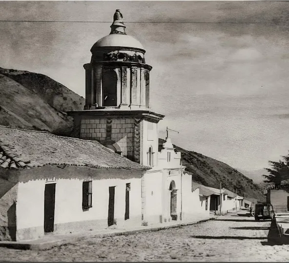 Fotografía antigua de la iglesia con su campanario de cúpula, la nave todavía baja y encalada, la calle sin asfaltar y un camión de los años treinta al fondo.
