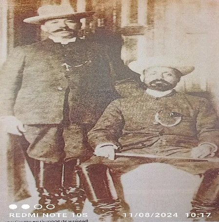 Dos militares de época, uno de pie con su fusil y otro sentado con bastón, en una fotografía sepia muy gastada.