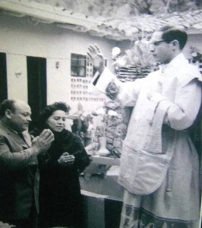 Un sacerdote joven, revestido, imparte la bendición a una pareja en el patio de una casa, en una fotografía antigua.