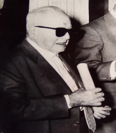 Un señor mayor de lentes oscuros y traje sostiene un pergamino enrollado.