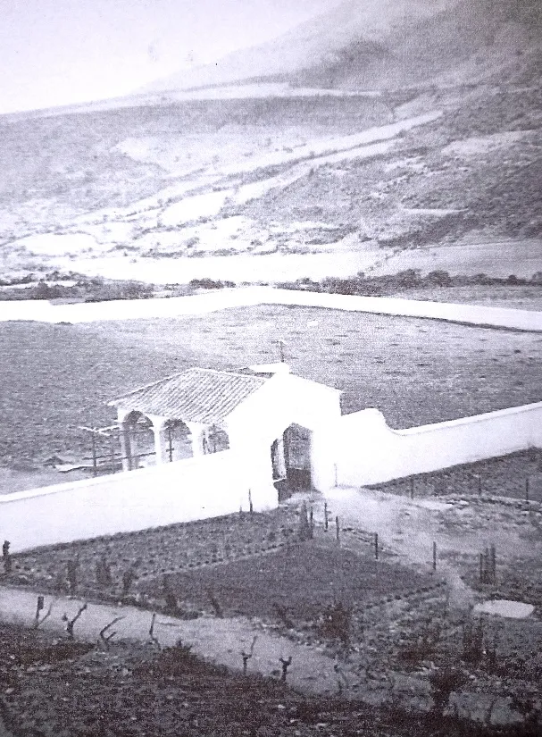 Fotografía antigua del cementerio: la capilla de tejas y su arco de entrada dentro de la tapia encalada, con los sembrados y el cerro alrededor.