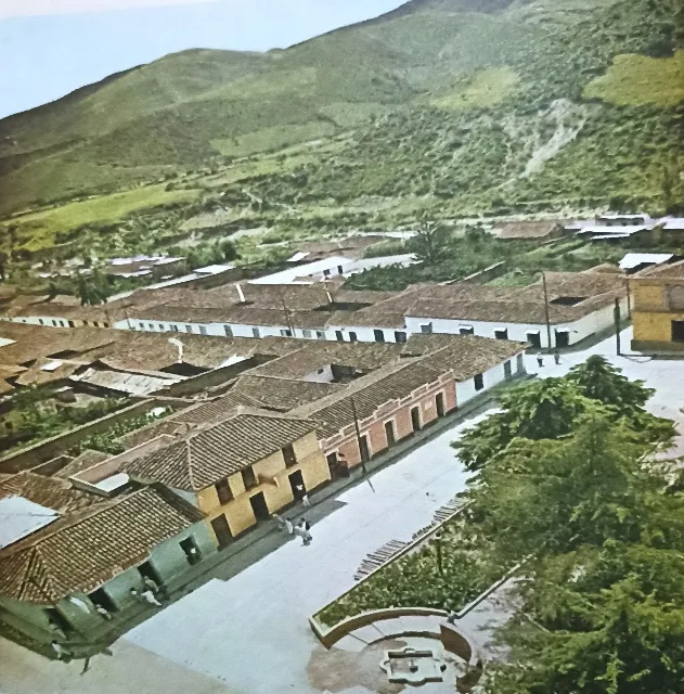 Vista aérea antigua y coloreada del pueblo: las hileras de casas de teja, la plaza con su glorieta y el cerro pelado detrás.