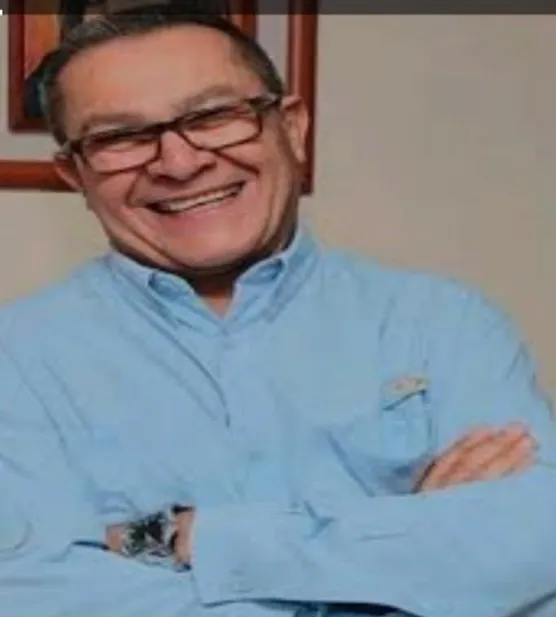 Retrato de un hombre sonriente, de lentes y camisa azul, con los brazos cruzados.