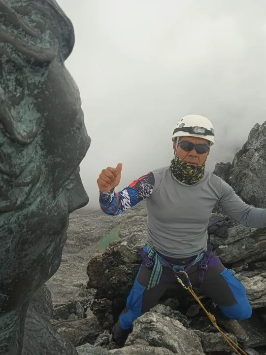 Un montañista con casco y arnés levanta el pulgar junto al busto de bronce del Libertador, en la cumbre entre la neblina.