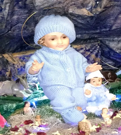 El Santo Niño vestido con gorro y ropa de lana azul, en el pesebre entre las flores y las figuras.