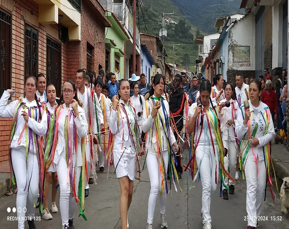 Comparsa de mujeres vestidas de blanco, con las cintas de colores al hombro y las maracas en la mano, bajando por la calle del pueblo.