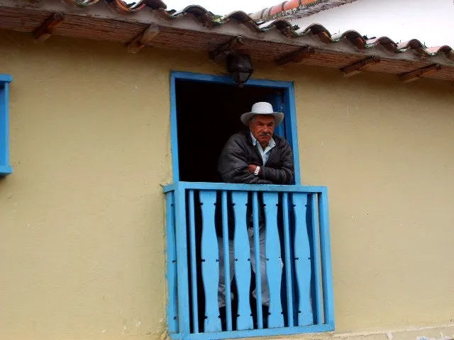 Un hombre de sombrero asomado a la ventana de balaustres azules de su casa de tapia, bajo el alero de tejas.