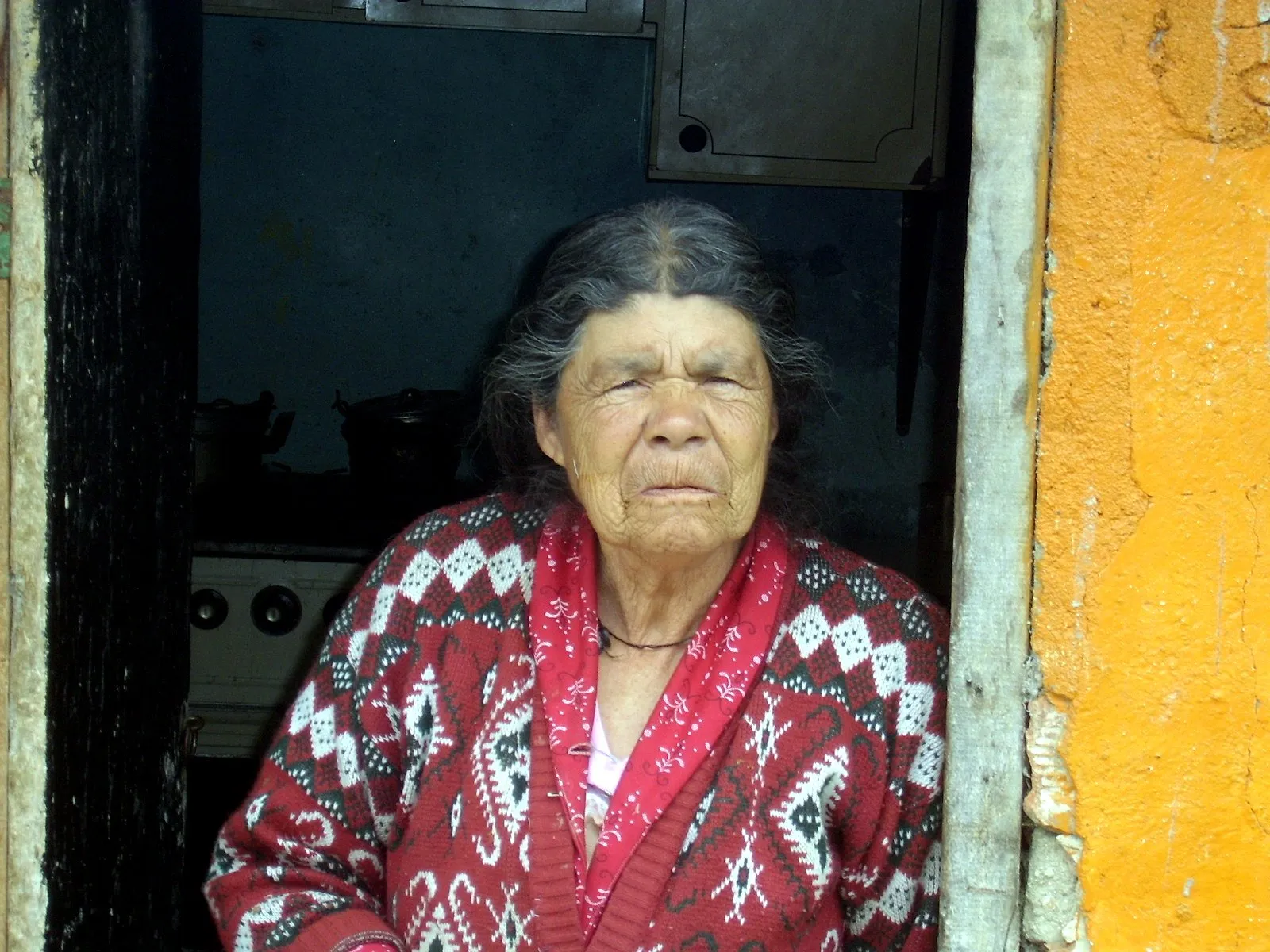 Una anciana en la puerta de su cocina, con el fogón detrás.