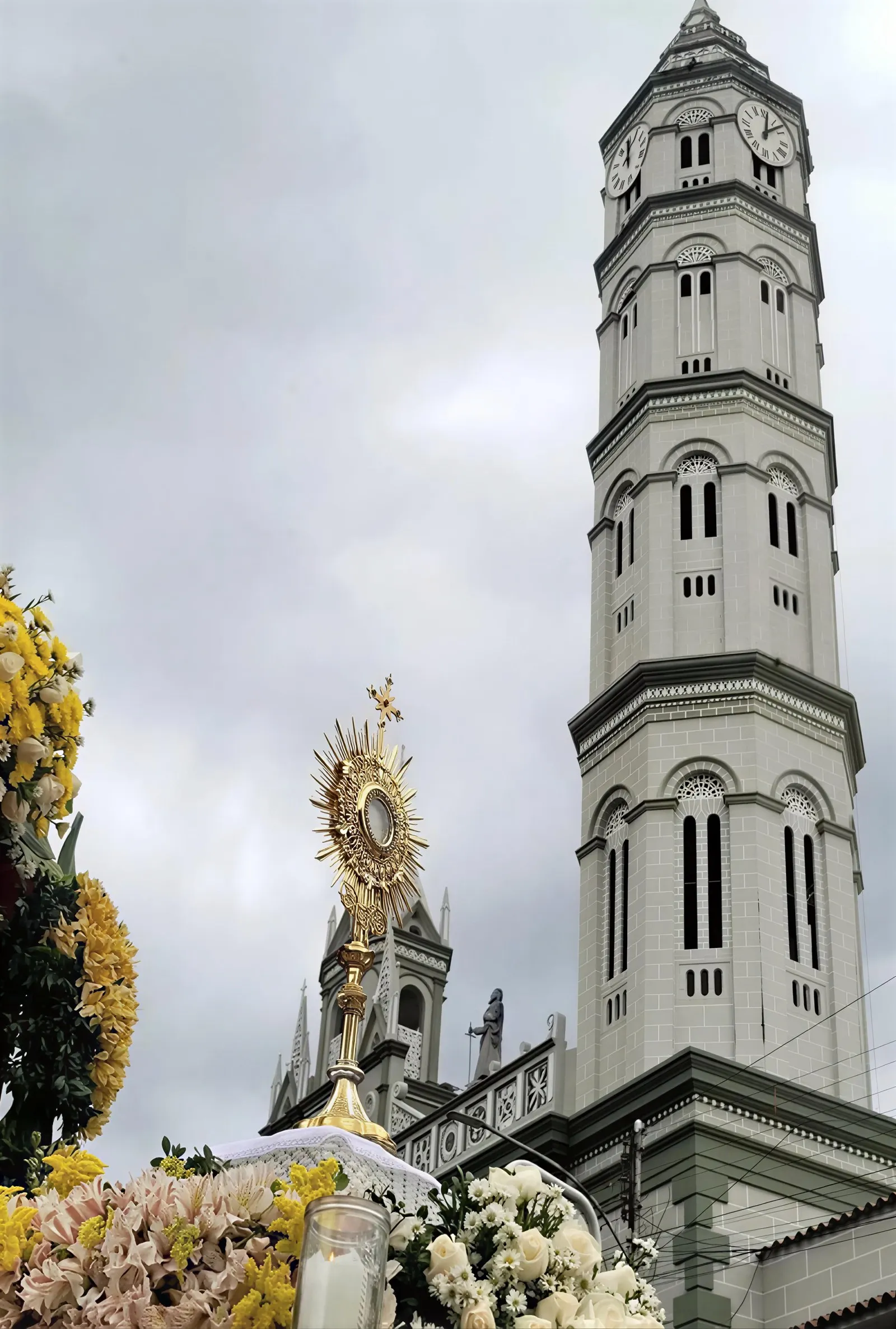 Una torre de campanario y una custodia dorada entre flores, durante una procesión.