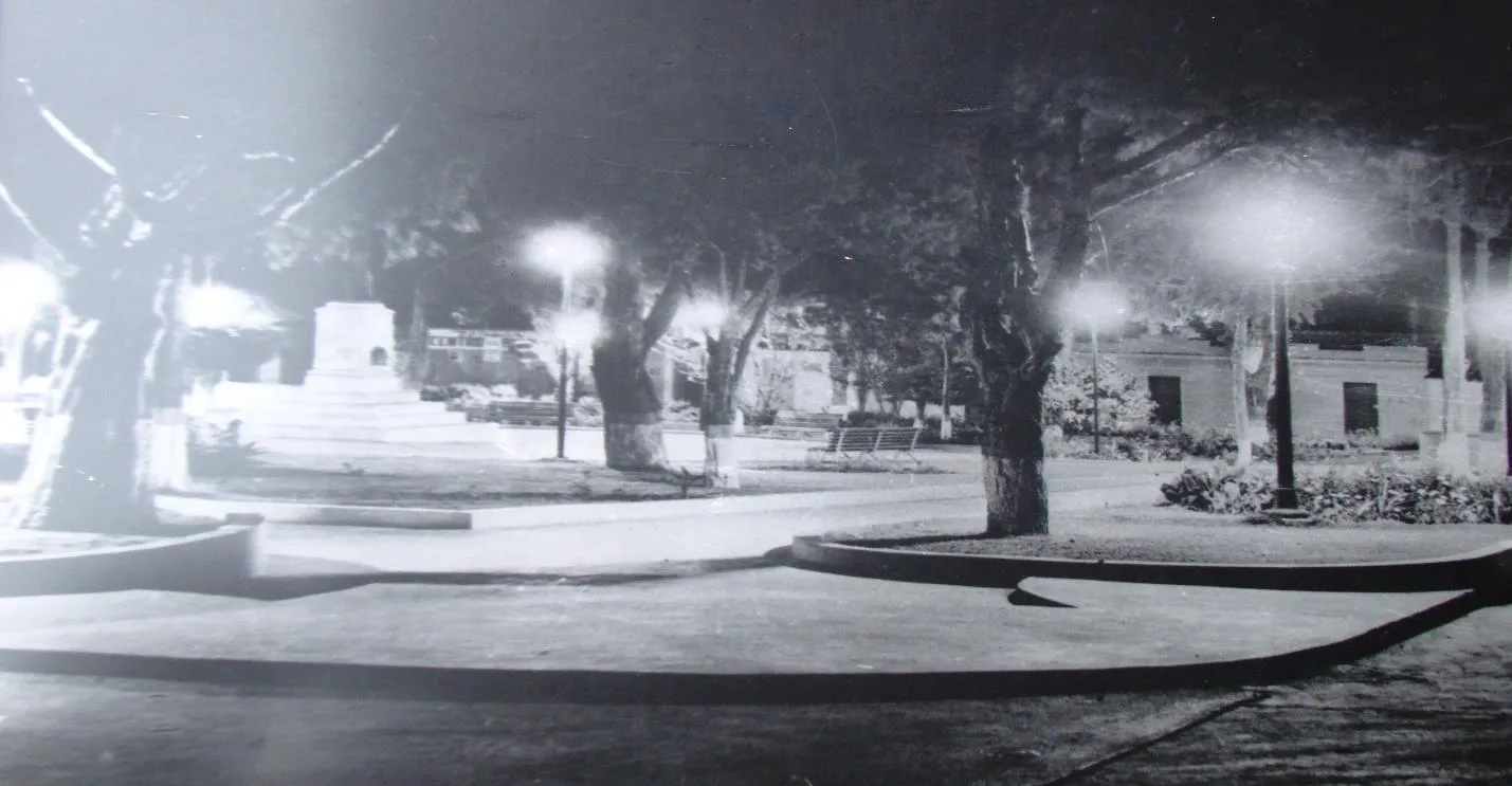 La plaza de noche, en una fotografía antigua: los faroles encendidos, los árboles y el pedestal del monumento.