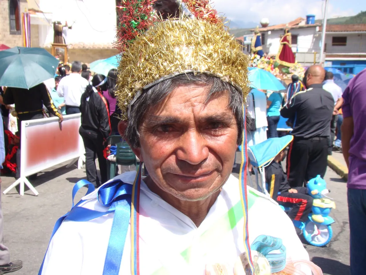 Giro de San Benito con la corona de oropel dorado y las cintas al hombro; detrás pasan las andas de la procesión.