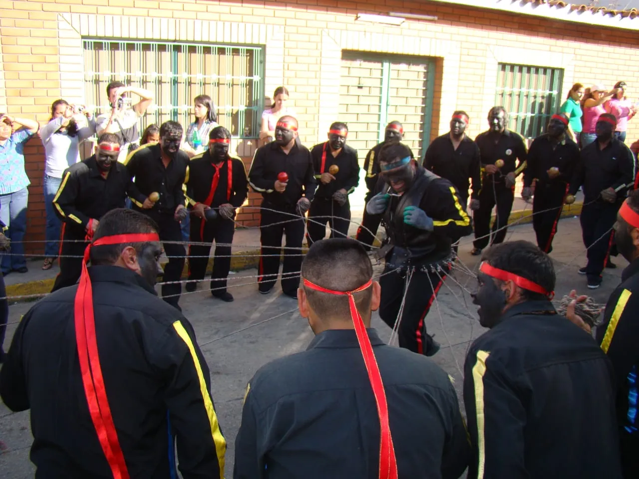 Los Negros de San Benito en rueda, de negro con franjas rojas y amarillas y el rostro tiznado, mientras uno baila enredado en los cordeles.