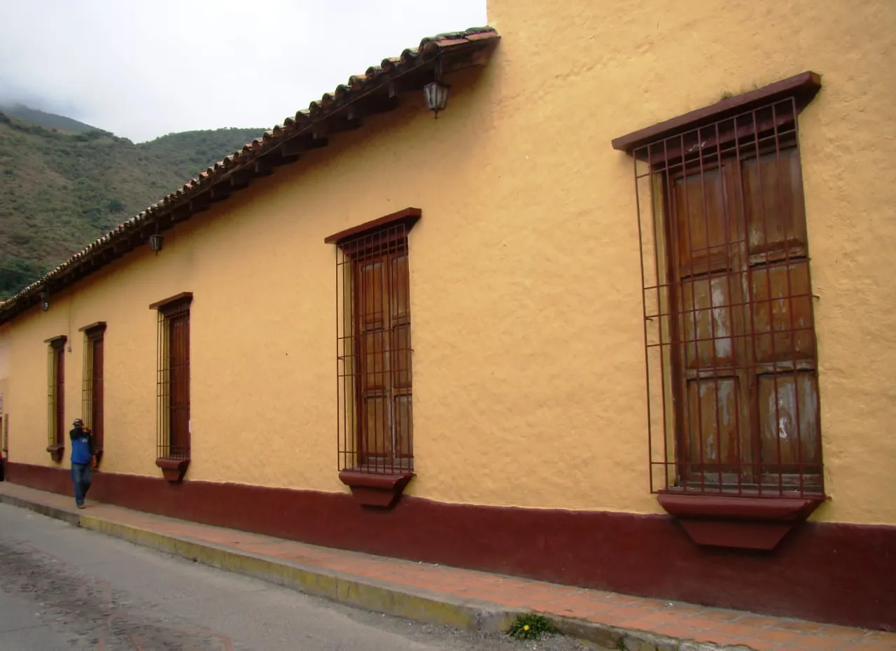 Casa de tapia encalada de ocre, con el zócalo vinotinto, las ventanas de madera enrejadas y el alero de tejas; al fondo el cerro.