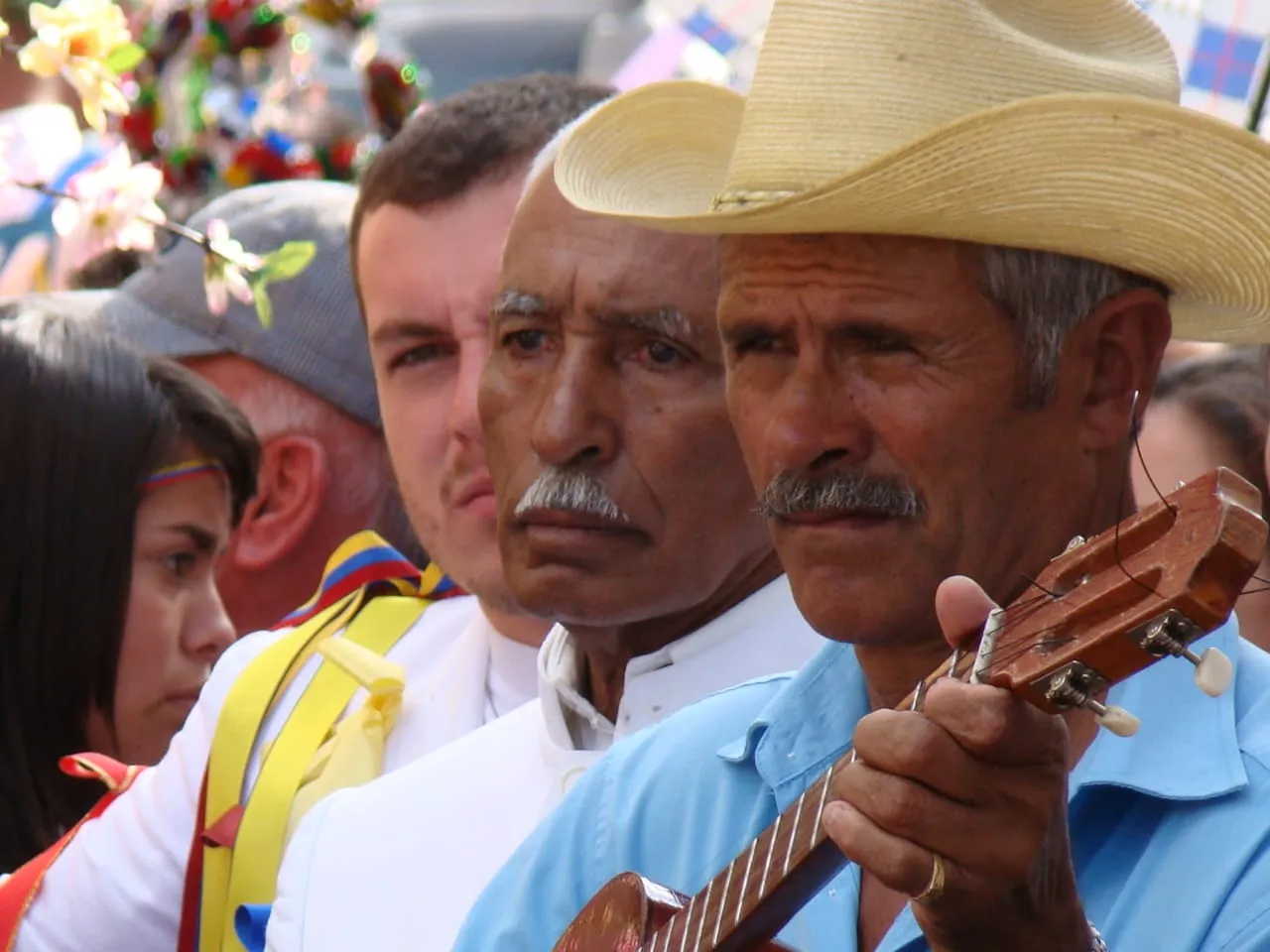 Músicos en la fiesta: uno rasga el cuatro y otros dos miran, con las cintas de los danzantes cruzándoles el hombro.