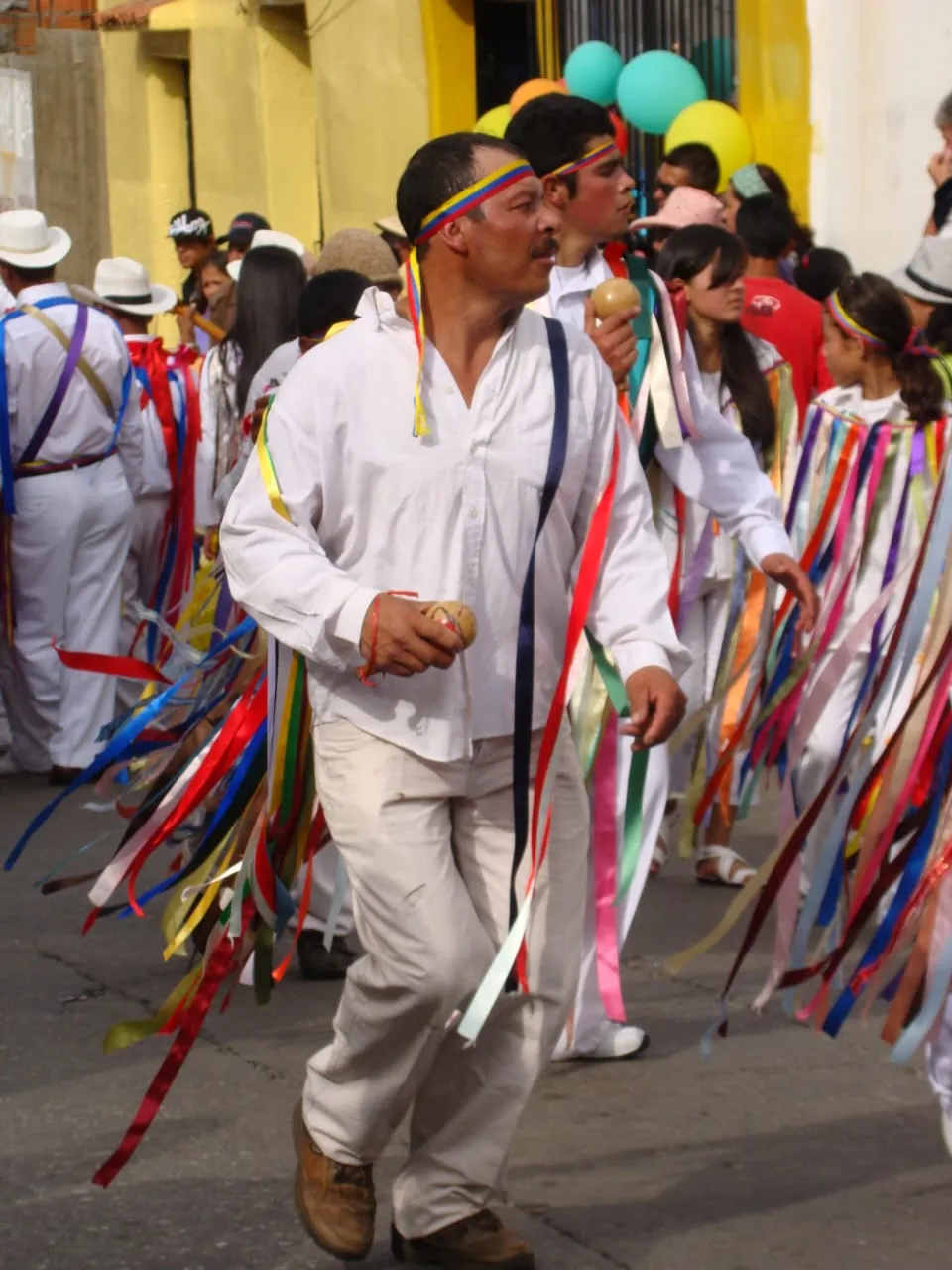 Danzante de blanco con la vincha tricolor y las cintas al vuelo, maraca en mano, bajando por la calle entre los demás.
